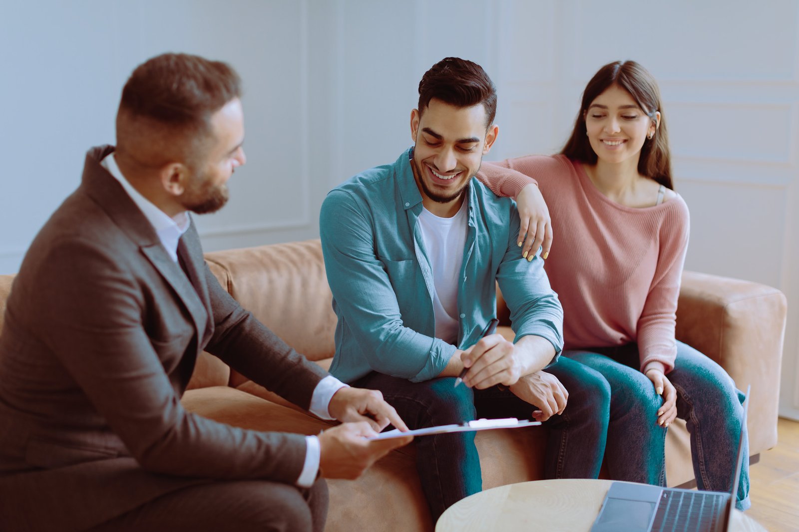 Happy Young Homeowners. Millennial Couple Buying New Private House, Financial Advisor In Suit Showing Contract, Smiling Man Signing Agreement Document, Sitting On The Couch At Office